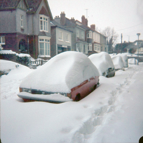 Car coch ar flaen y llun wedi'i orchuddio ag eira, ac yna llinell o geir wedi'u parcio i bob golwg y tu allan i res o dai (hefyd wedi'u gorchuddio gan eira).