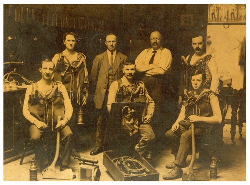 Ffotograff Sepia o saith dyn - pump yn ffitio i fyny mewn offer anadlu hen ffasiwn, a dau yn sefyll y tu ôl i'r grŵp yn edrych yn awdurdodol. Mae offer mwyngloddio amrywiol yn lledaenu’r llawr.